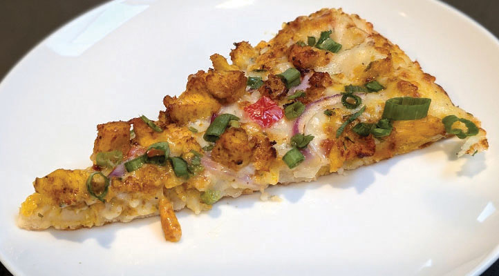 A slice of curry pizza adorned with anchari chicken and many vegetables.